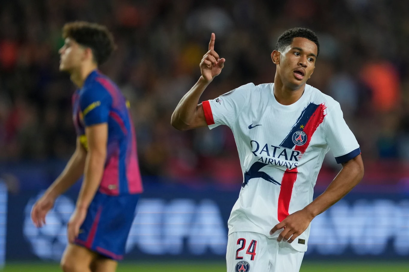 Piłkarz w białym stroju Paris Saint-Germain unosi palec do góry po zdobytej bramce, za nim drugi zawodnik w granatowo-bordowym stroju z opuszczoną głową, rozmyte tło stadionu umożliwia skupienie uwagi na emocjach obu graczy.