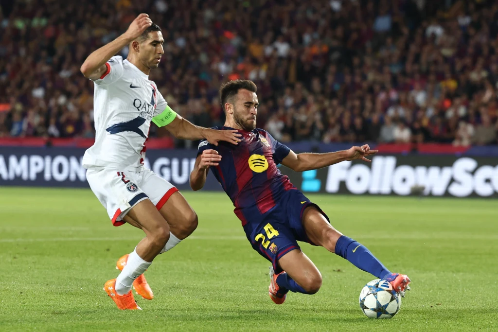 Dwóch piłkarzy rywalizuje o piłkę podczas meczu, jeden z zawodników wykonuje wślizg, a w tle widać trybuny pełne kibiców.