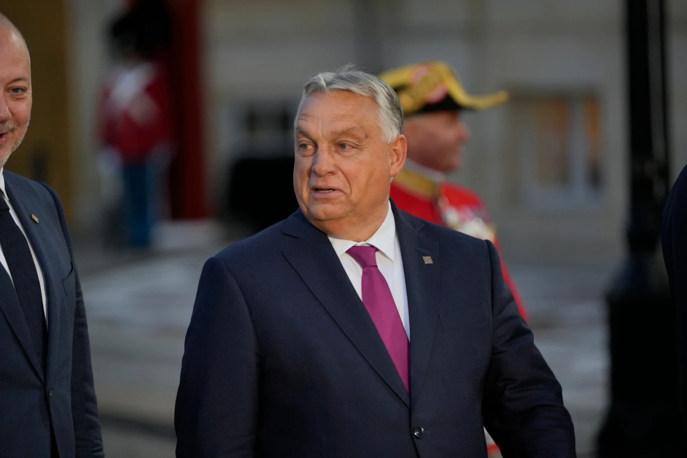 Premier Węgier Viktor Orban Mężczyzna w garniturze i fioletowym krawacie, patrzący w bok, w tle żołnierz w paradnym mundurze oraz inne postaci w rozmytym tle.