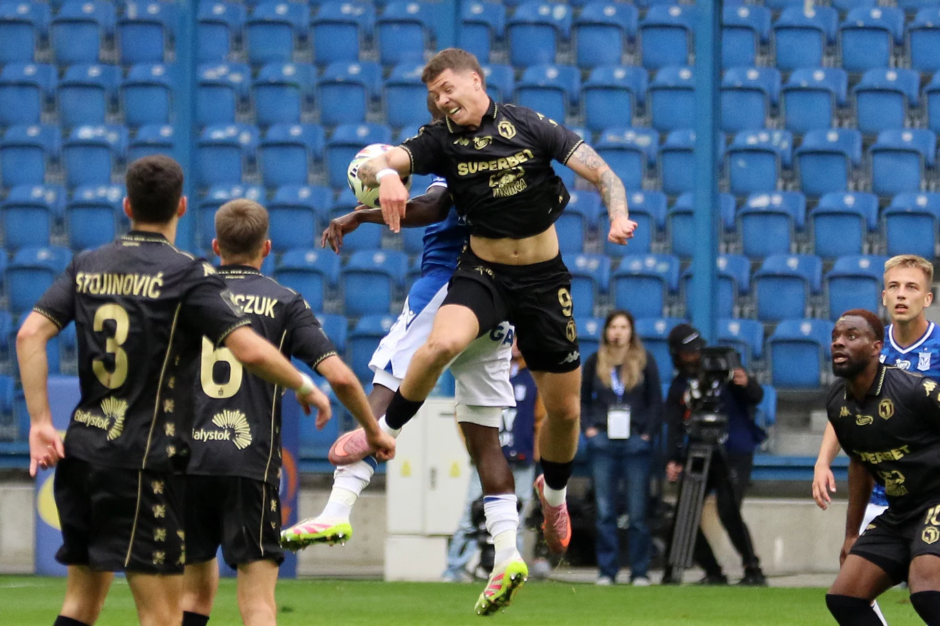 Piłkarze walczą o piłkę głową w trakcie meczu na stadionie, otoczeni przez innych zawodników i pustymi niebieskimi trybunami w tle.