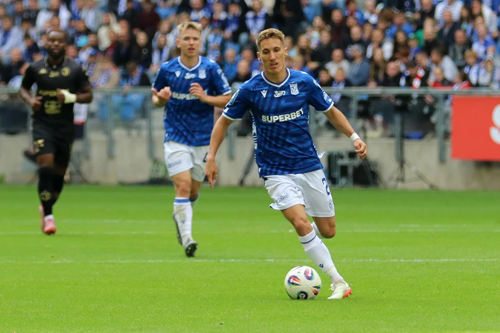 Joel Pereira, Lech Poznań Piłkarz w niebieskiej koszulce drybluje piłkę po murawie stadionu, w tle widoczny kolejny zawodnik oraz kibice na trybunach.