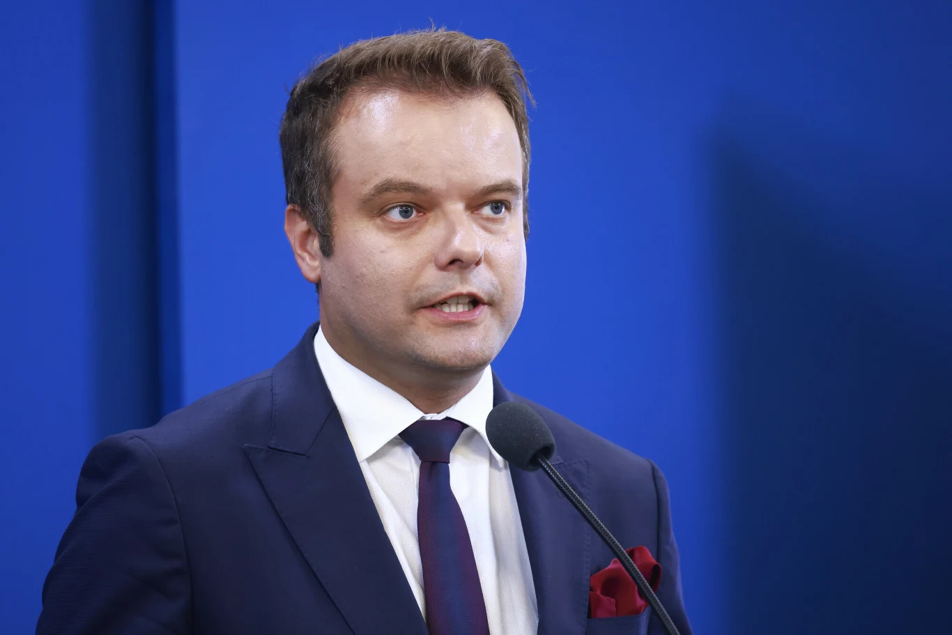 Rzecznik PiS Rafał Bochenek Mężczyzna w garniturze i krawacie przemawiający do mikrofonu na tle niebieskiej ściany.