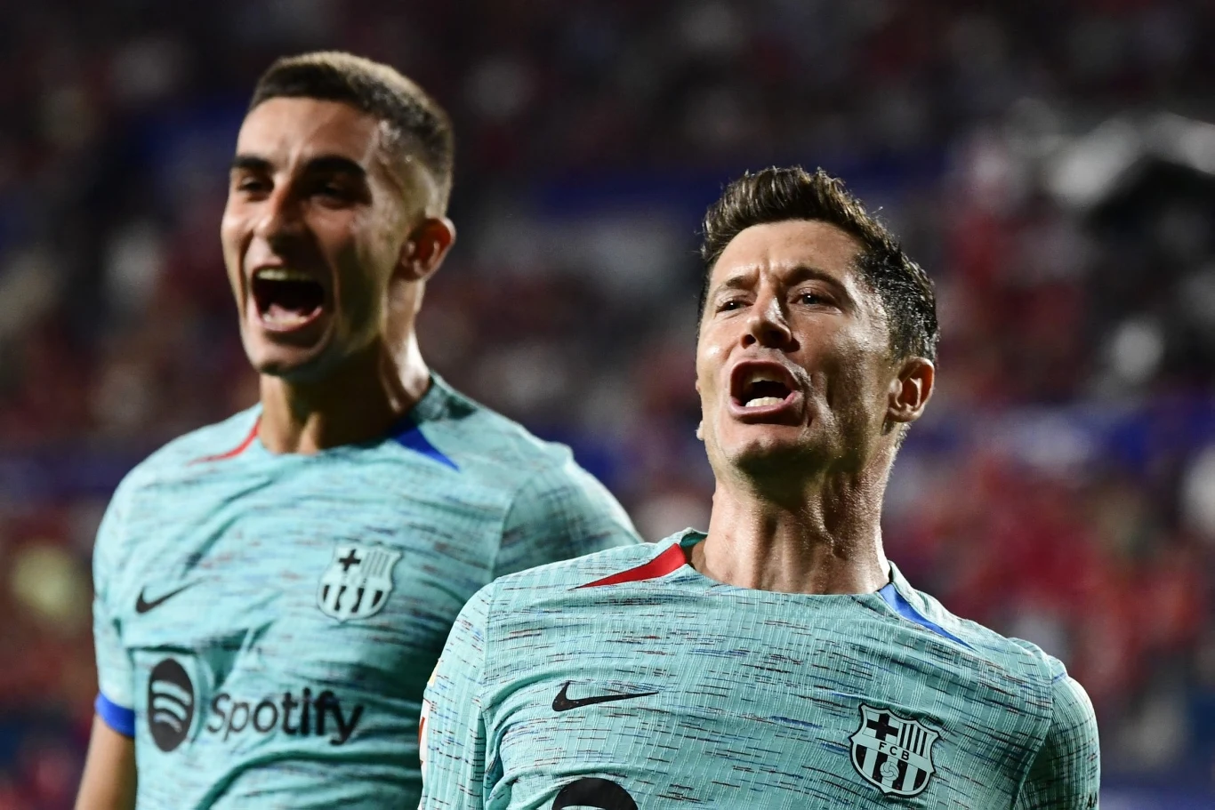 Dwóch piłkarzy FC Barcelony w jasnozielonych strojach świętuje zdobycie gola na tle rozmazanej publiczności stadionu.