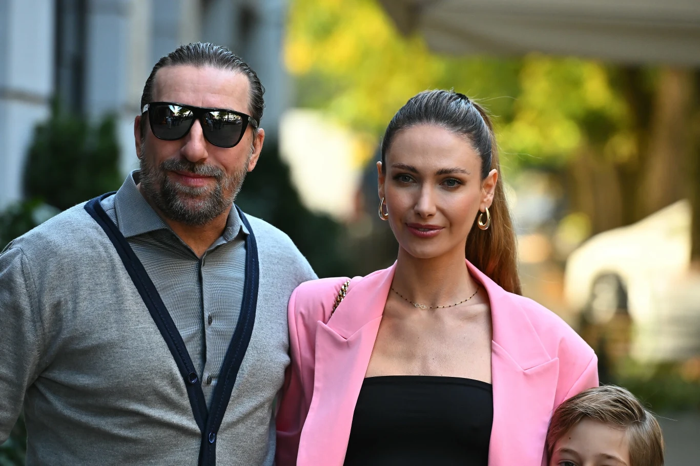 Mężczyzna w okularach przeciwsłonecznych i kobieta w eleganckim różowym żakiecie stoją razem na tle miejskiego parku, obok widoczna głowa dziecka.