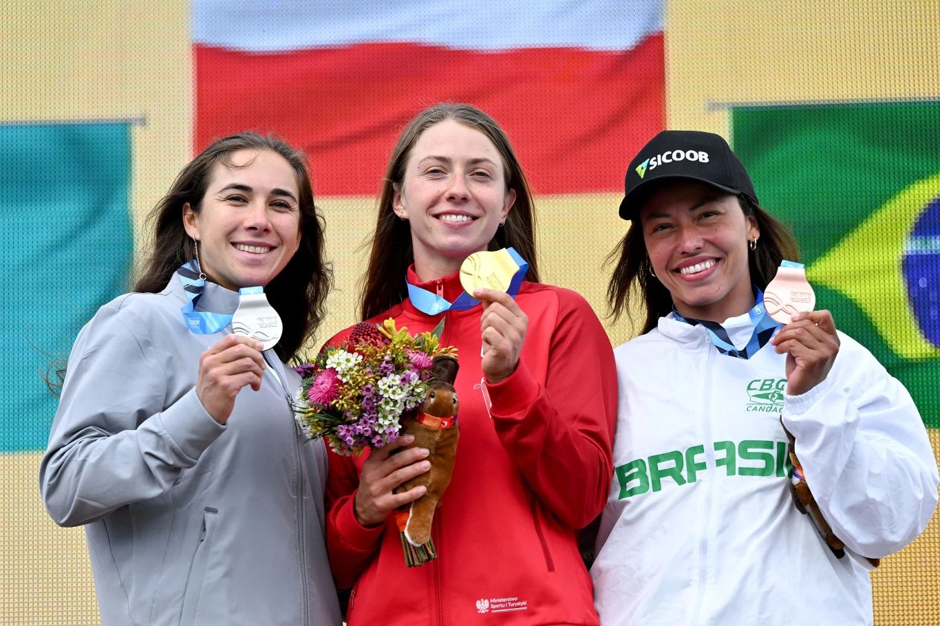 Trzy kobiety stoją na podium z medalami na szyi, środkowa trzyma bukiet kwiatów, w tle widoczne są flagi Polski i Brazylii, wszystkie uśmiechają się i prezentują swoje medale.