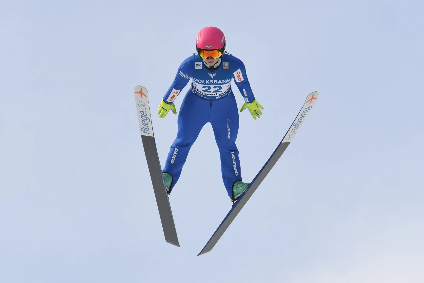 Zawodnik w niebieskim kombinezonie i różowym kasku wykonujący skok narciarski w locie, dłonie ułożone wzdłuż ciała, narty szeroko rozłożone w powietrzu na tle jasnego nieba.