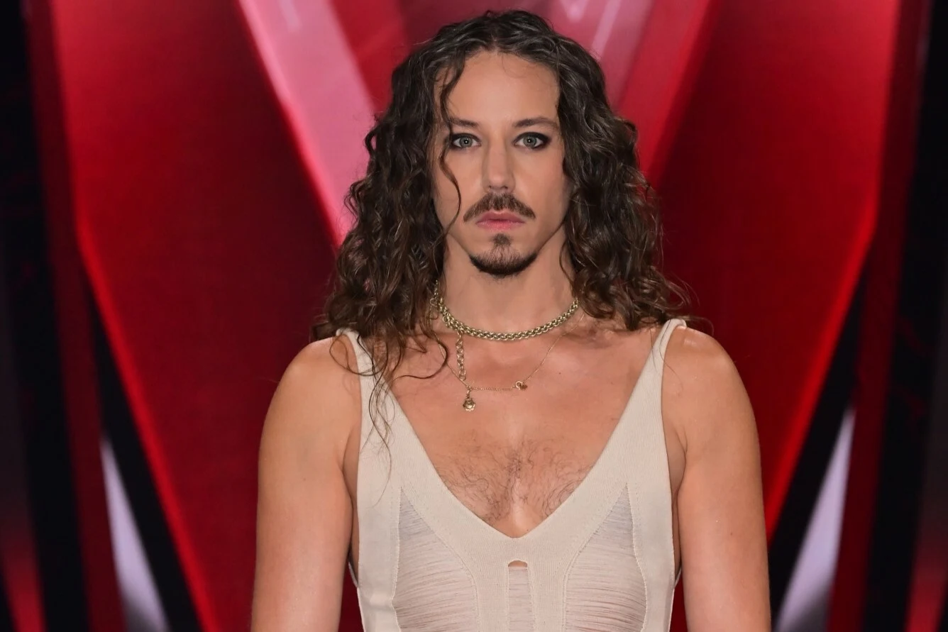 Michał Szpak Mężczyzna z długimi kręconymi włosami, brodą i wąsami, ubrany w jasną, dopasowaną sukienkę z głębokim dekoltem, na szyi złoty łańcuszek, w tle czerwono-czarna scenografia z wyraźnym podświetleniem.