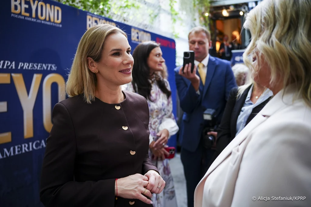 Pierwsza Dama z polskimi businesswoman w Nowym Jorku. (źródło zdjęcia: Kancelaria Prezydenta RP/ autor: Alicja Stefaniuk) Oficjalne spotkanie kilku osób ubranych elegancko, rozmowa kobiet na pierwszym planie, tło z banerem promocyjnym w barwach granatowo-złotych, w tle mężczyzna z aparatem fotograficznym.