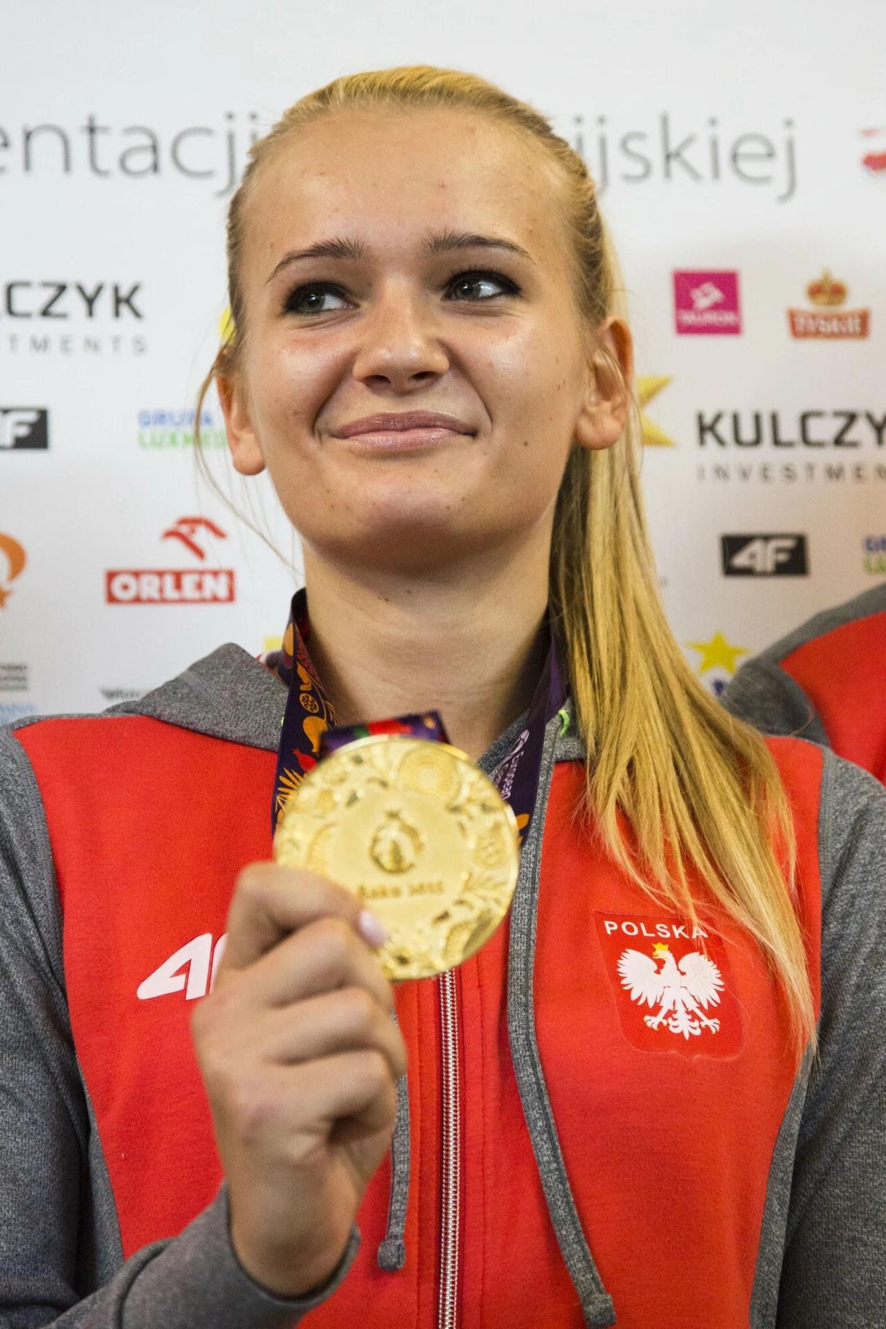 Młoda kobieta w czerwonym stroju reprezentacyjnym Polski, trzymająca złoty medal i uśmiechająca się, na tle planszy z logotypami sponsorów.
