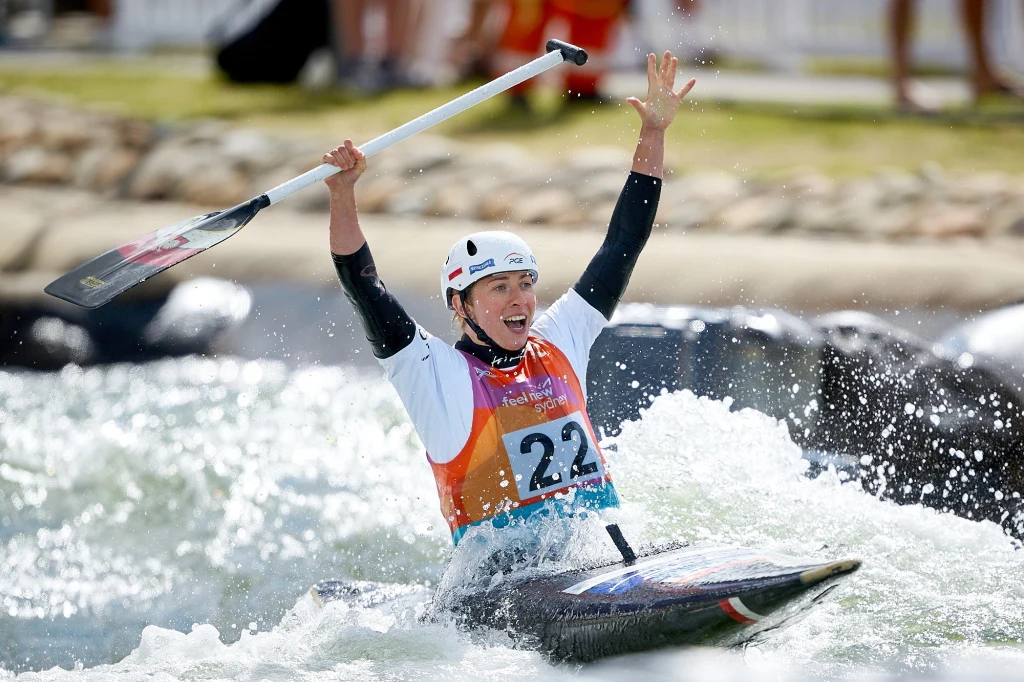Klaudia Zwolińska Uśmiechnięty kajakarz w kasku i kamizelce startowej oznaczonej numerem 22 unosi ręce oraz wiosło w geście radości podczas płynięcia przez wzburzoną wodę, w tle rozmyte sylwetki kibiców przy brzegu.