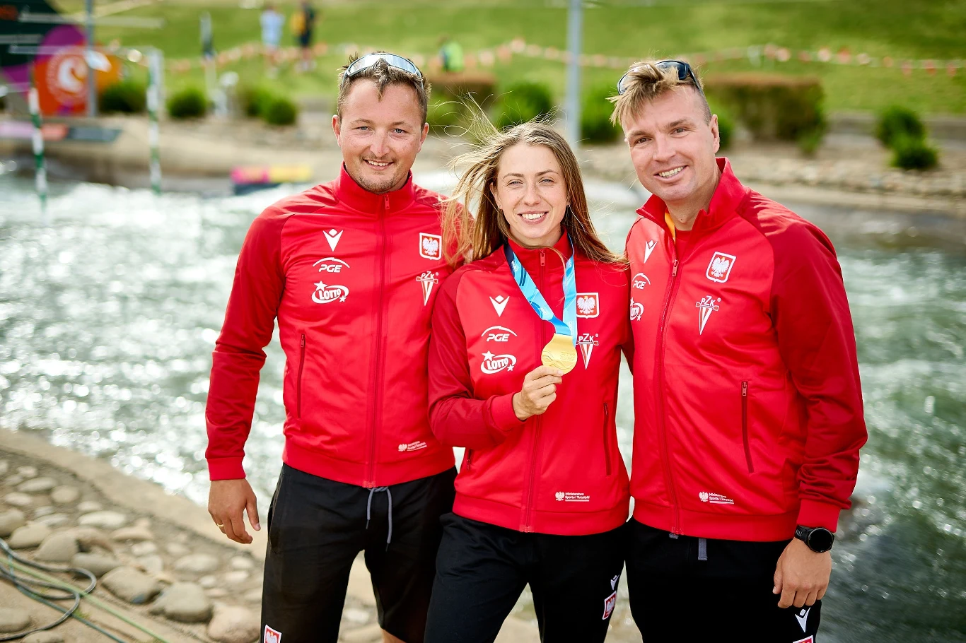 Trzech sportowców w czerwonych kurtkach z polskim godłem pozuje nad wodnym torem, środkowa osoba trzyma złoty medal i uśmiecha się, wszyscy prezentują radość i dumę z osiągnięcia.