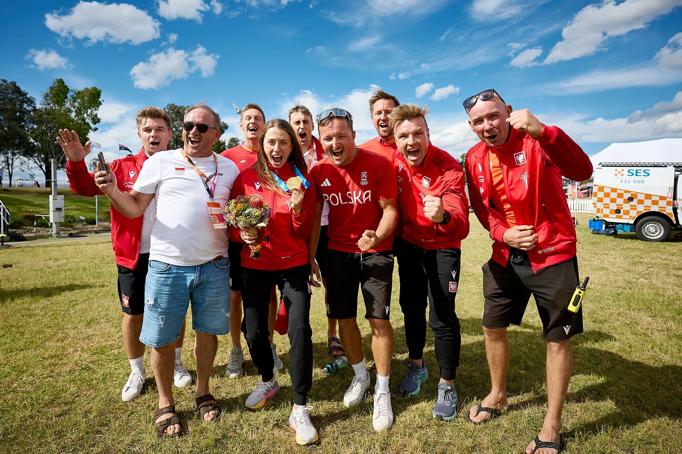 Grupa uśmiechniętych sportowców i trenerów ubranych w czerwone stroje z napisem Polska, stojących na trawie pod gołym niebem i celebrujących sukces, jedna kobieta trzyma bukiet kwiatów, w tle widać karetkę oraz błękitne niebo z chmurami.
