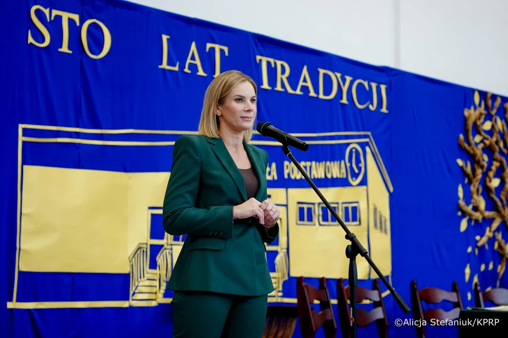 Marta Nawrocka podczas w uroczystościach 100–lecia nadania Szkole Podstawowej w Liwie im. Stefana Batorego(źródło: Kancelaria Prezydenta RP, autora: Alicja Stefaniuk) Kobieta w zielonym garniturze przemawia do mikrofonu na tle niebieskiej dekoracji z napisem 'Sto lat tradycji' i rysunkiem budynku szkoły podstawowej, z ustawionymi obok krzesłami.