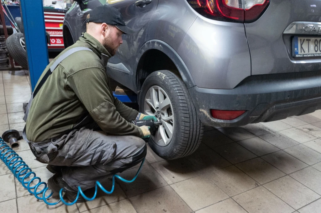 Ile kosztuje wymiana opon w 2025 roku? Mechanik pracuje przy wymianie koła w srebrnym samochodzie podniesionym na podnośniku, używając narzędzi pneumatycznych, w tle widoczne wyposażenie warsztatu.