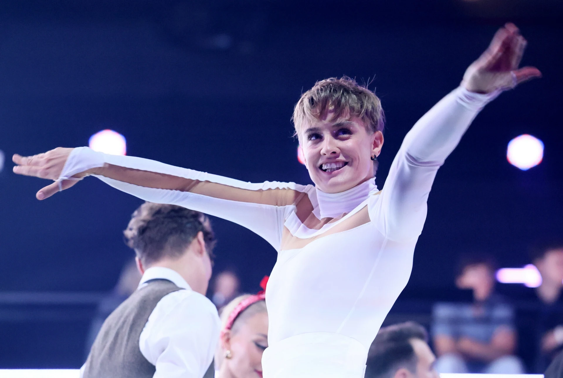 Sportowiec w białym stroju z rozłożonymi ramionami uśmiecha się, w tle widzowie oraz inni uczestnicy wydarzenia sportowego.