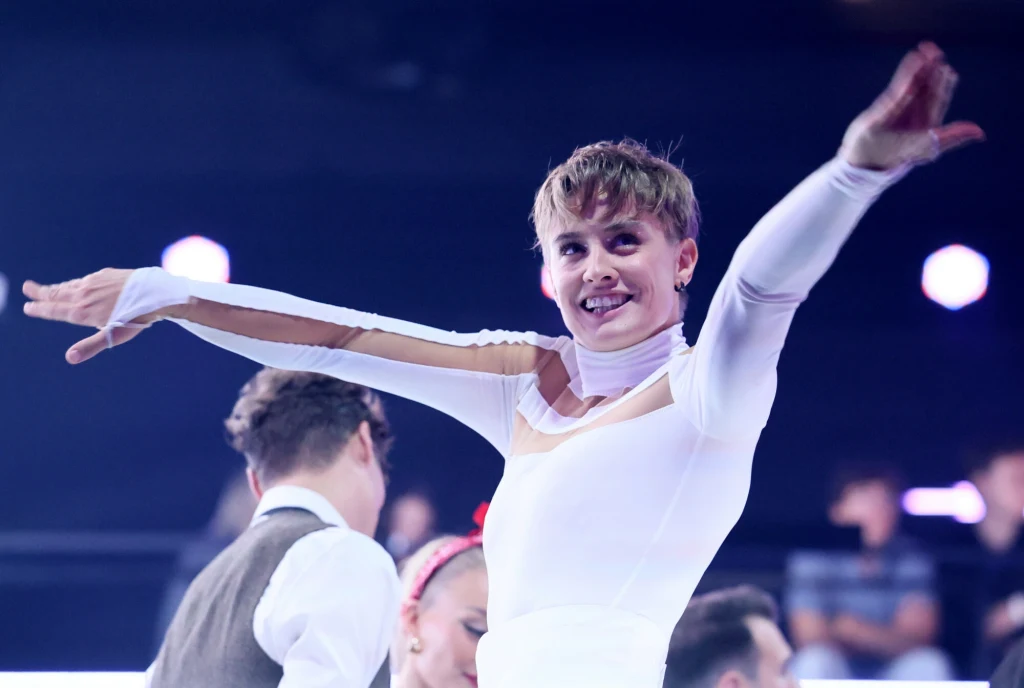 Katarzyna Zillmann w programie "Taniec z Gwiazdami" Sportowiec w białym stroju z rozłożonymi ramionami uśmiecha się, w tle widzowie oraz inni uczestnicy wydarzenia sportowego.