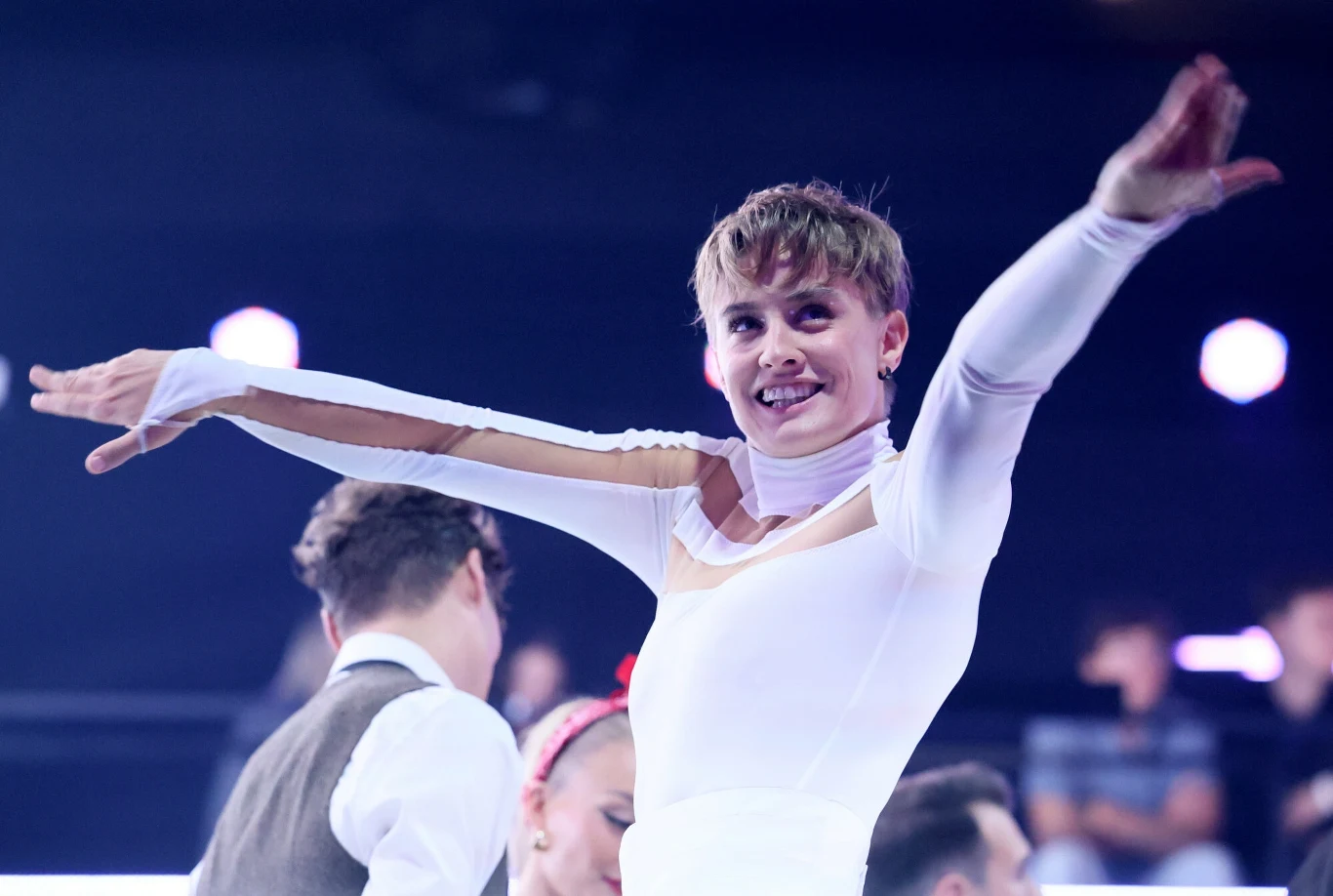 Katarzyna Zillmann w programie "Taniec z Gwiazdami" Sportowiec w białym stroju z rozłożonymi ramionami uśmiecha się, w tle widzowie oraz inni uczestnicy wydarzenia sportowego.