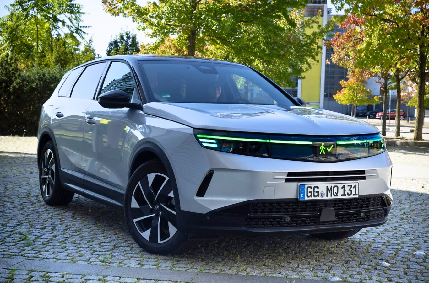 Nowoczesny srebrny SUV ustawiony na kamiennej kostce, otoczony zielonymi drzewami i budynkami w tle. Charakterystyczne zielone podświetlenie z przodu samochodu i niemieckie tablice rejestracyjne.