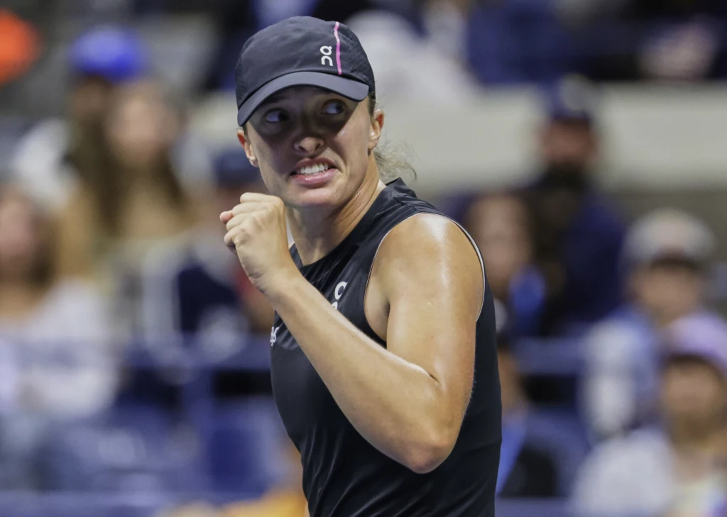 Iga Świątek Zdeterminowana tenisistka w czarnej czapce i stroju sportowym świętuje zwycięstwo z zaciśniętą pięścią na tle widowni.