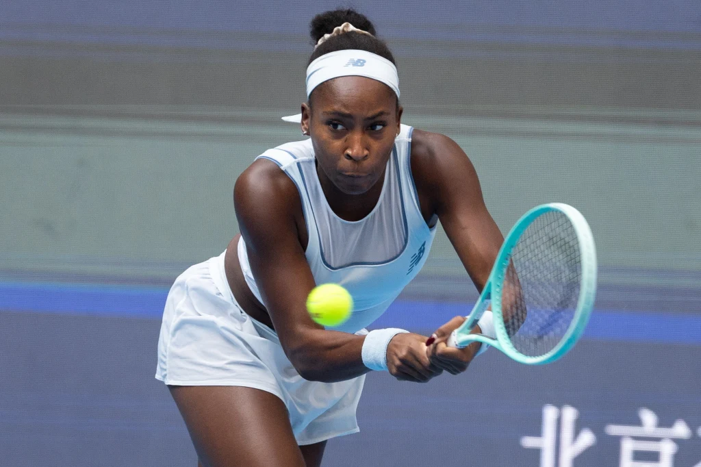 Sportowiec w białym stroju tenisowym skupiony podczas wykonania uderzenia dwuręcznego backhandu, żółta piłka tenisowa tuż przy rakiecie, niebieskie tło kortu.