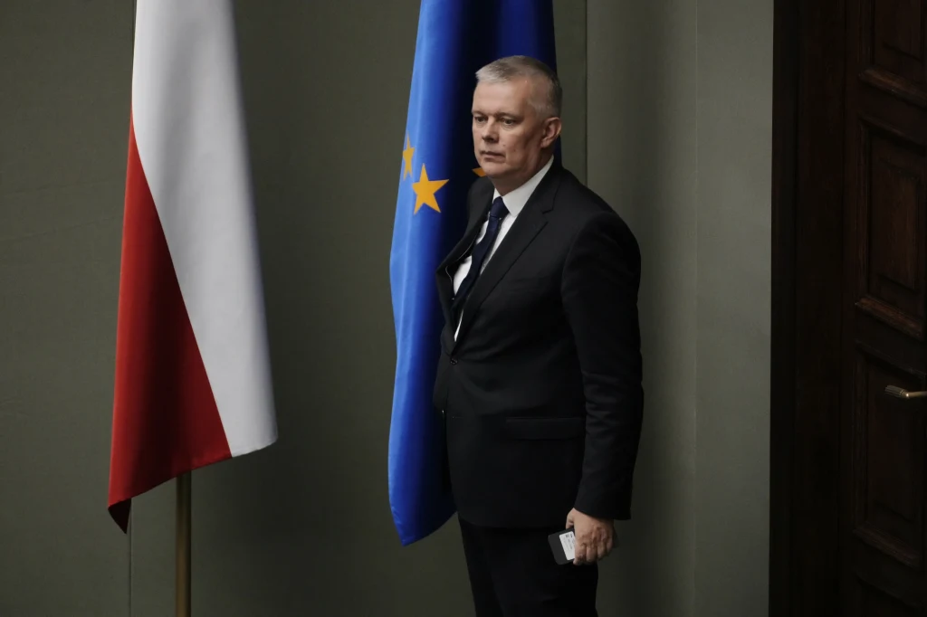 Minister koordynator służb specjalnych Tomasz Siemoniak Mężczyzna w garniturze stoi obok flagi Polski i flagi Unii Europejskiej na tle ciemnej ściany wewnątrz budynku, trzymając w dłoni dokument.