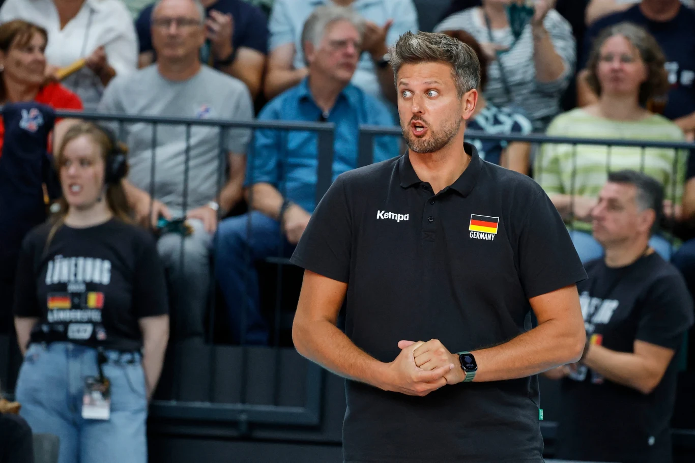 Michał Winiarski jest selekcjonerem reprezentacji Niemiec Trener drużyny narodowej Niemiec w siatkówce mężczyzn w czarnej koszulce z godłem Niemiec i napisem Germany, stojący na tle publiczności podczas meczu, z wyraźnie skupioną miną.