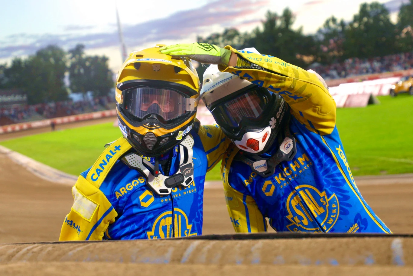 Dwóch żużlowców w żółto-niebieskich kombinezonach i kaskach pozuje razem na torze, otoczeni kibicami na tle stadionu.