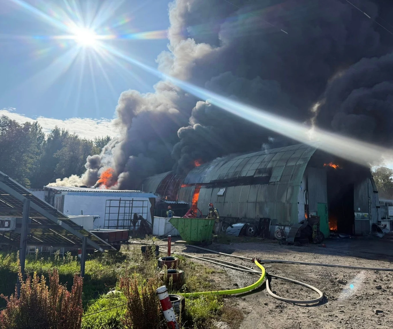 Intensywny pożar hali magazynowej, z której unoszą się gęste, czarne kłęby dymu, sylwetki strażaków próbujących opanować ogień, w tle widać jasne słońce oraz otoczenie przemysłowe z porozrzucanymi sprzętami i szlaką.