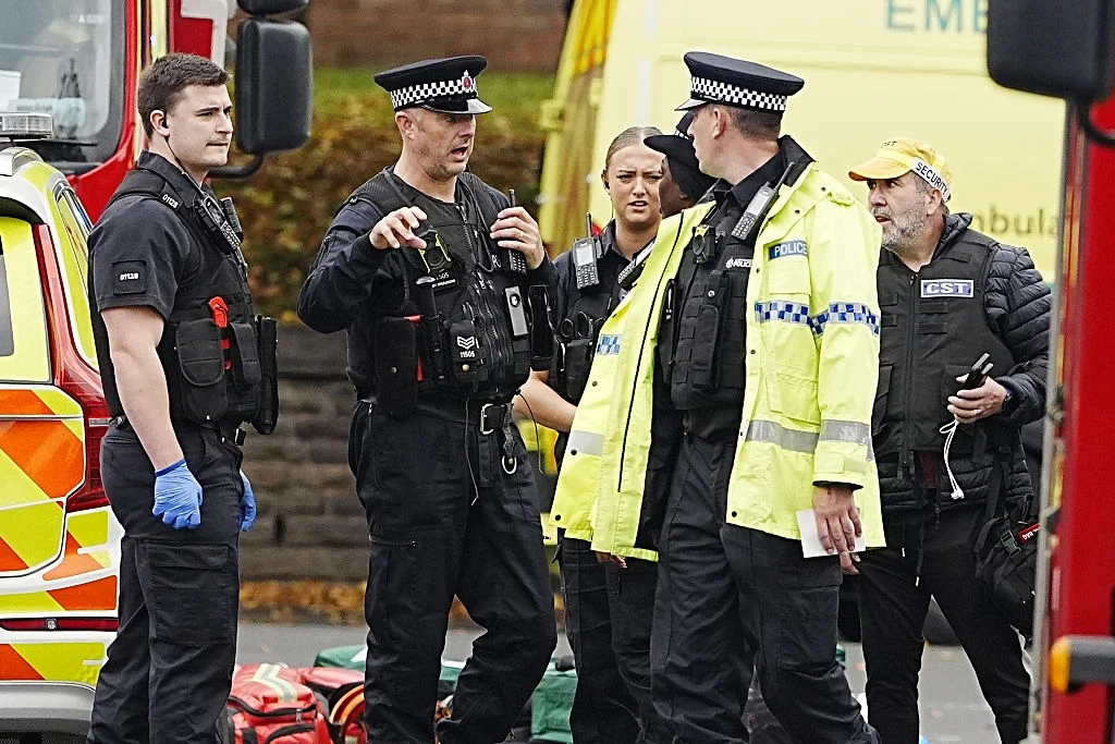 Atak na synagogę w Manchesterze Grupa policjantów i osób ratowniczych w mundurach i kamizelkach odblaskowych stoi w pobliżu pojazdów służb ratunkowych, prowadząc rozmowę i koordynując działania.