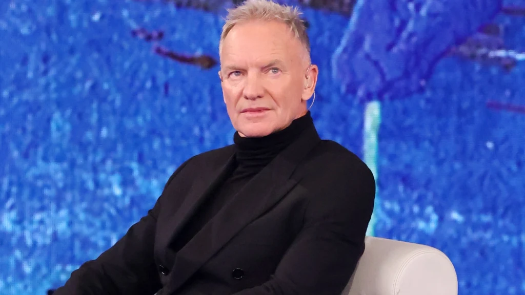 Sting Mężczyzna w czarnym garniturze i czarnym golfie siedzi na jasnym fotelu na tle niebieskiego obrazu w tle.