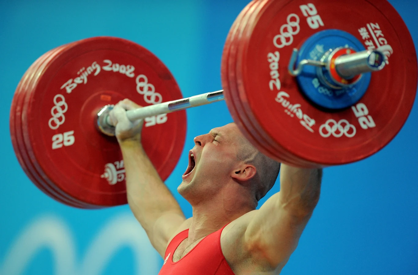 Zawodnik w czerwonym stroju sportowym unosi ciężar olimpijski nad głową, wyrażając wysiłek i emocje podczas rywalizacji na tle niebieskiego, oficjalnego tła igrzysk olimpijskich.