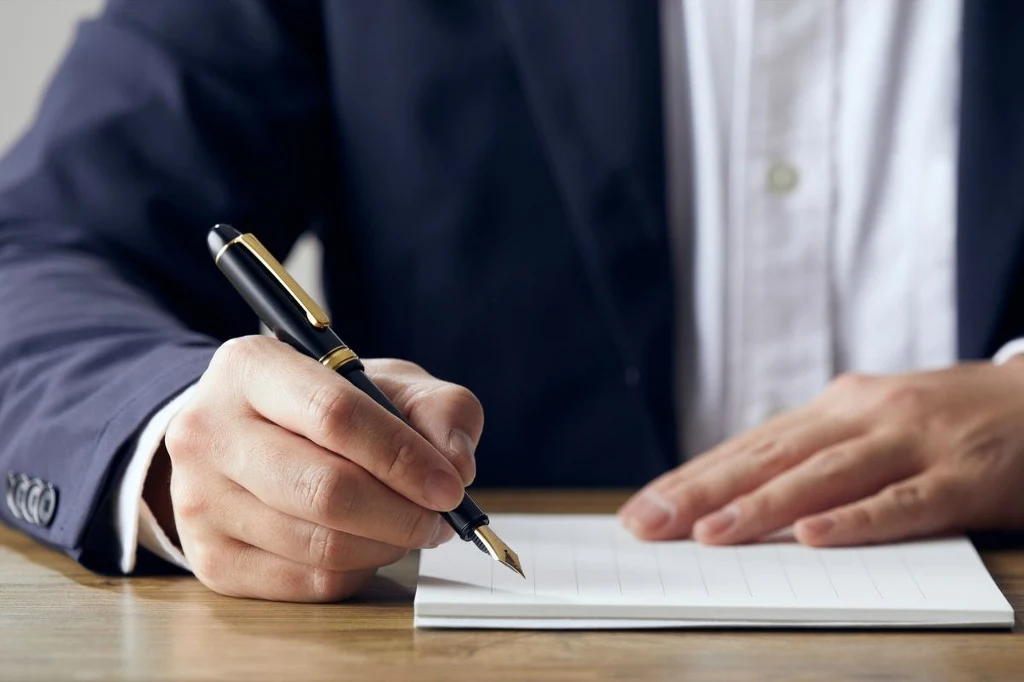 Wieczne pióra były niegdyś symbolem luksusu Mężczyzna w garniturze pisze piórem na białej kartce papieru, siedząc przy drewnianym stole.