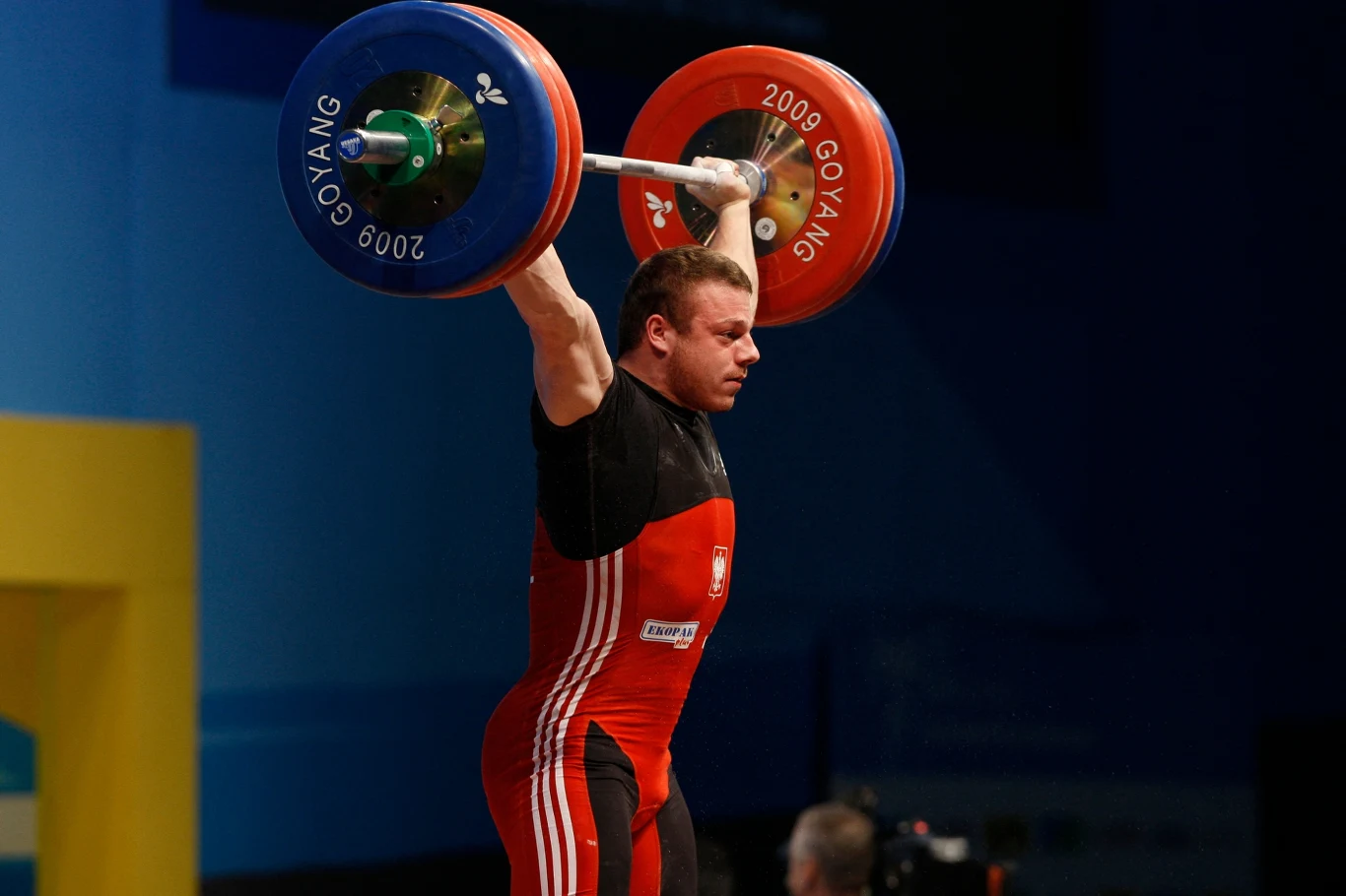 Sportowiec unoszący nad głową ciężar podczas zawodów podnoszenia ciężarów, ubrany w czerwony strój startowy, skupiony na utrzymaniu sztangi z dużymi obciążnikami.