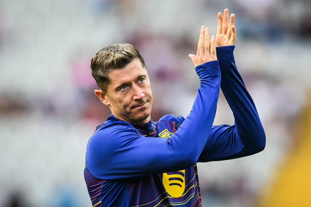 Robert Lewandowski Piłkarz w sportowej bluzie bije brawo, jego dłonie uniesione są na wysokości głowy, tło rozmyte, skupienie na mimice twarzy sportowca.