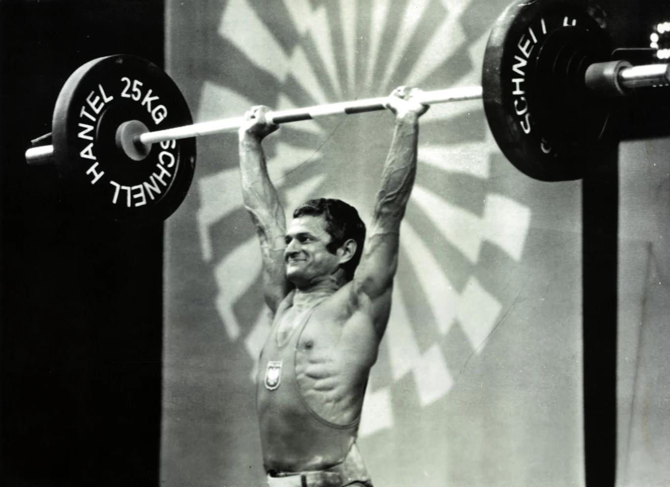 Sportowiec w stroju prezentującym godło narodowe podczas podnoszenia sztangi nad głową na arenie sportowej, w tle geometryczne wzory rozświetlające scenę.