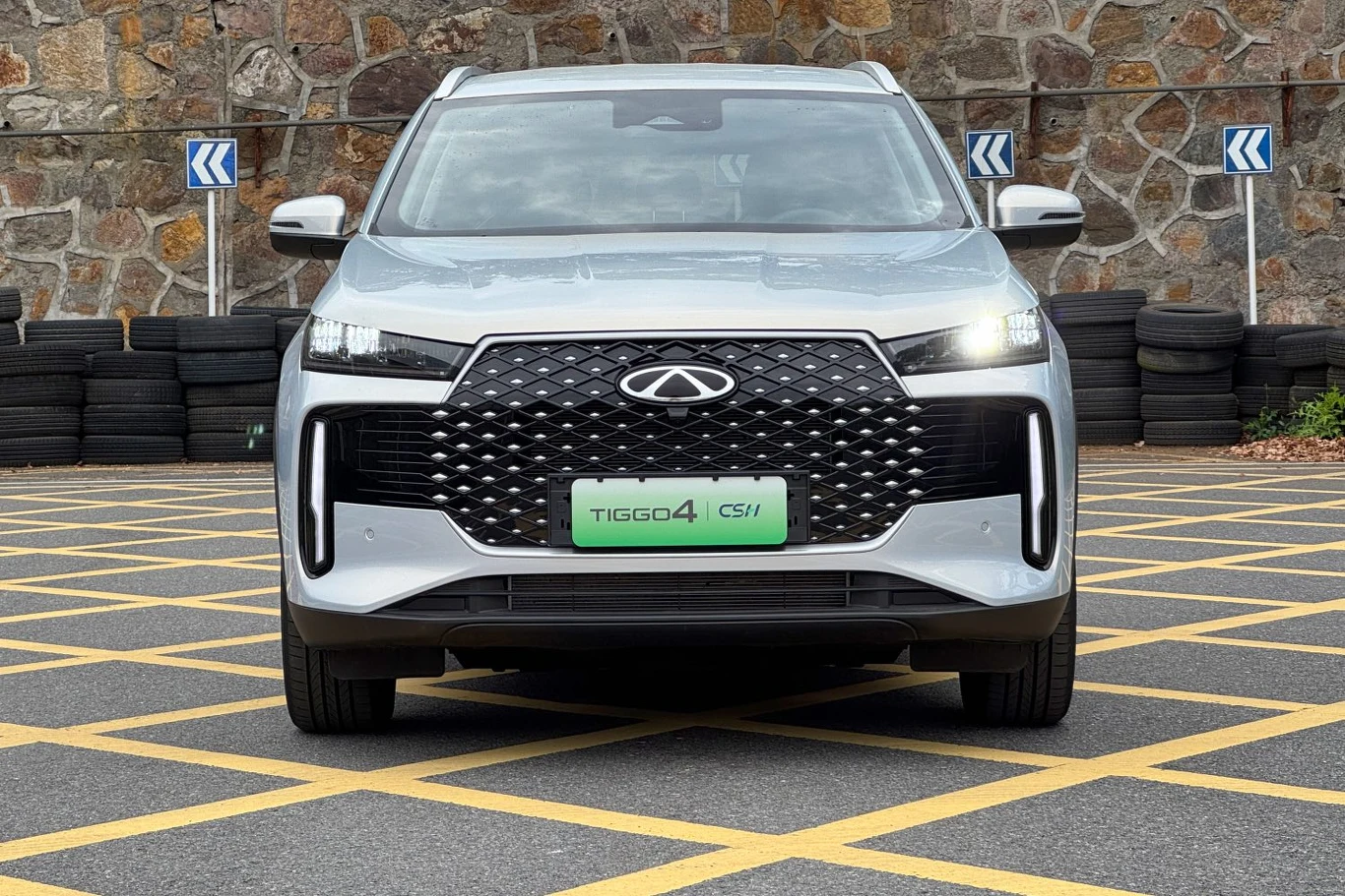 Srebrny samochód osobowy marki Chery Tiggo 4, stojący na żółto oznakowanym parkingu przed kamiennym murem z oponami ułożonymi w tle. Widok z przodu ukazuje nowoczesny grill i reflektory LED.