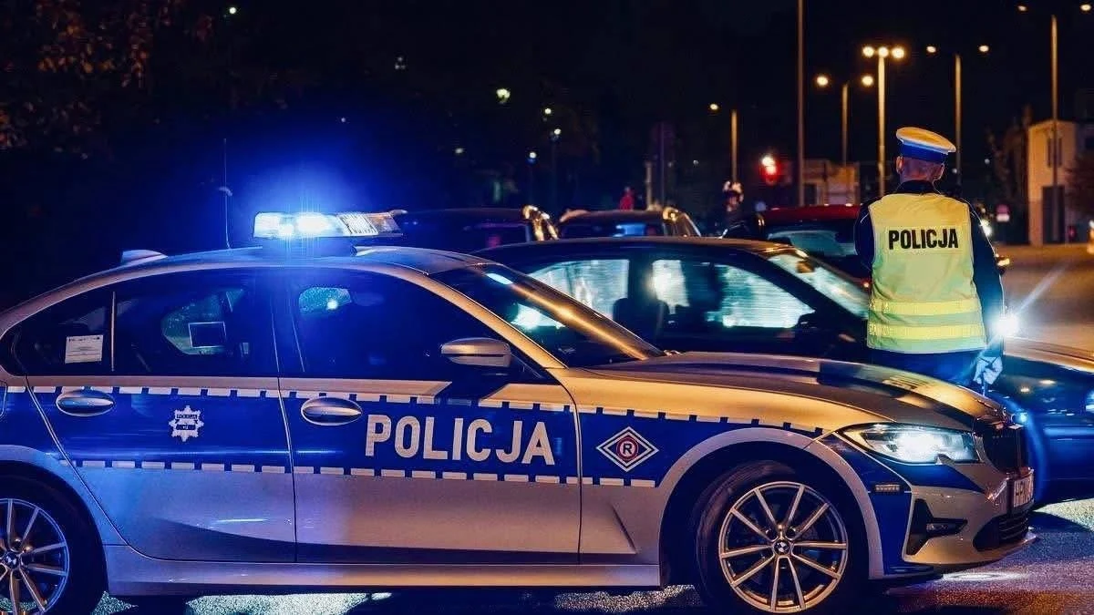 Do tragicznych wydarzeń doszło w nocy w hotelu w Wolsztynie/ Zdj. ilustracyjne Radiowóz policyjny z włączonymi światłami stoi na ulicy nocą, obok policjant w żółtej kamizelce w pobliżu drugiego samochodu, w tle rozświetlone latarnie i niewyraźne sylwetki osób.
