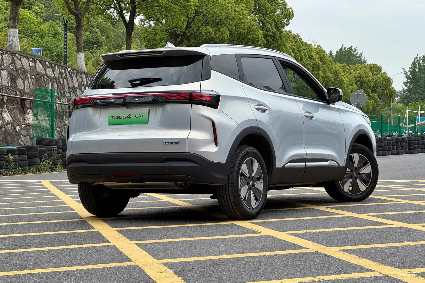 Srebrny samochód typu SUV marki Chery Tiggo 4, zaparkowany na oznaczonych, żółtych liniach na ulicy, w tle widoczny kamienny mur oraz zieleń drzew.