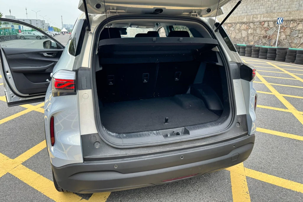 Podłoga bagażnika nie jest idealnie płaska – widać, gdzie pod nią znajduje się akumulator Otwarty bagażnik srebrnego samochodu osobowego zaparkowanego na żółtych liniach na parkingu, tylne drzwi otwarte, widoczny fragment wnętrza pojazdu oraz tylna kanapa.