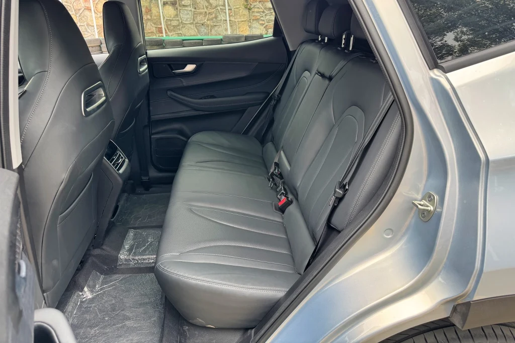 Jak na swoje gabaryty Tiggo 4 może się pochwalić całkiem przestronnym wnętrzem Tylny rząd siedzeń samochodu z czarną, skórzaną tapicerką, szerokimi fotelami i wyraźnie widocznymi pasami bezpieczeństwa, w tle fragment okna oraz drzwi samochodu otwarty na oścież.