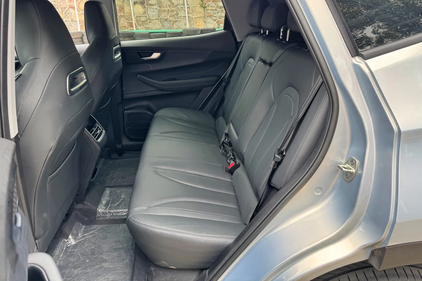 Tylny rząd siedzeń samochodu z czarną, skórzaną tapicerką, szerokimi fotelami i wyraźnie widocznymi pasami bezpieczeństwa, w tle fragment okna oraz drzwi samochodu otwarty na oścież.