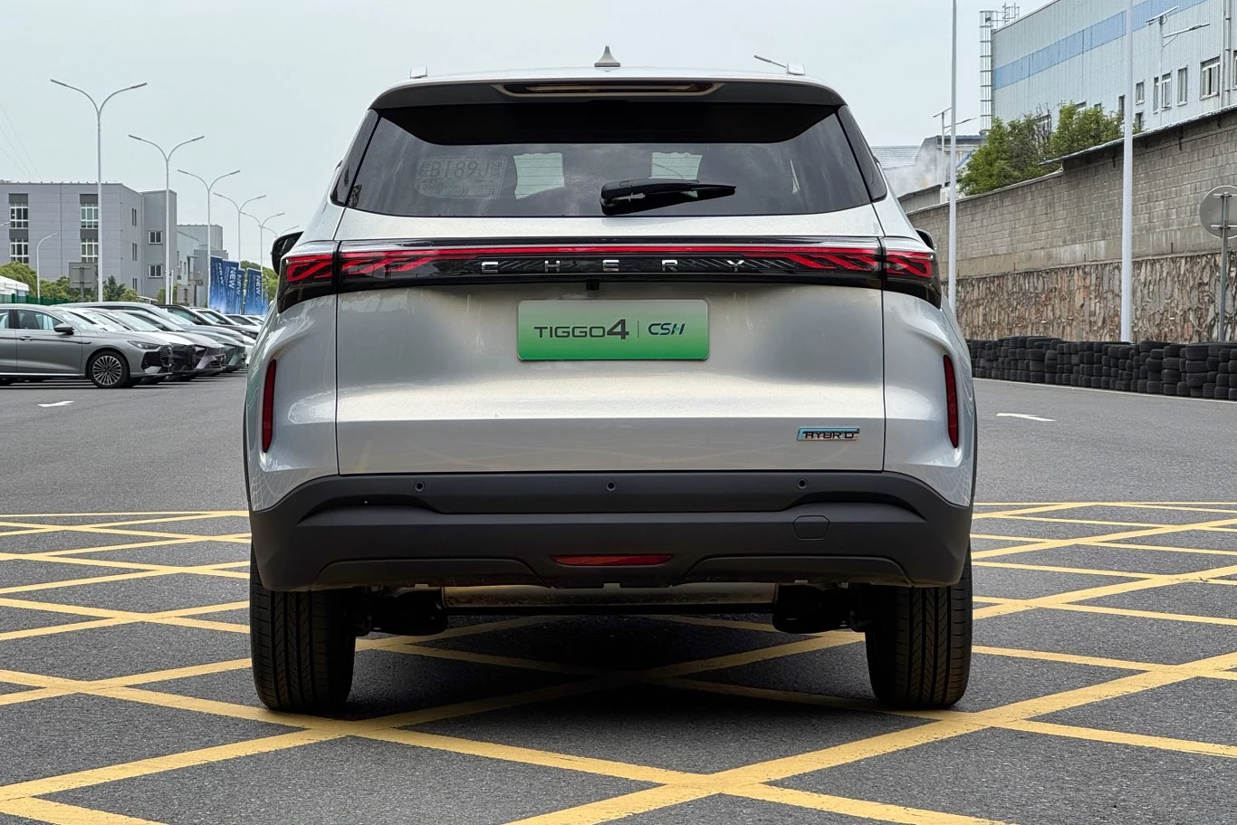 Srebrny samochód SUV Chery Tiggo 4 Pro widziany od tyłu, zaparkowany na oznaczonym miejscu parkingowym, w tle widoczne inne samochody oraz zabudowania przemysłowe.