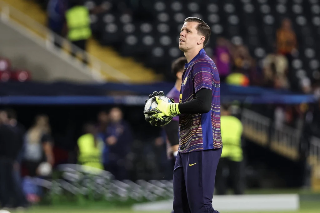 Wojciech Szczęsny Bramkarz w stroju sportowym stoi na murawie stadionu, trzymając piłkę do gry w dłoniach, w tle widoczni są inni ludzie oraz trybuny z pustymi miejscami.