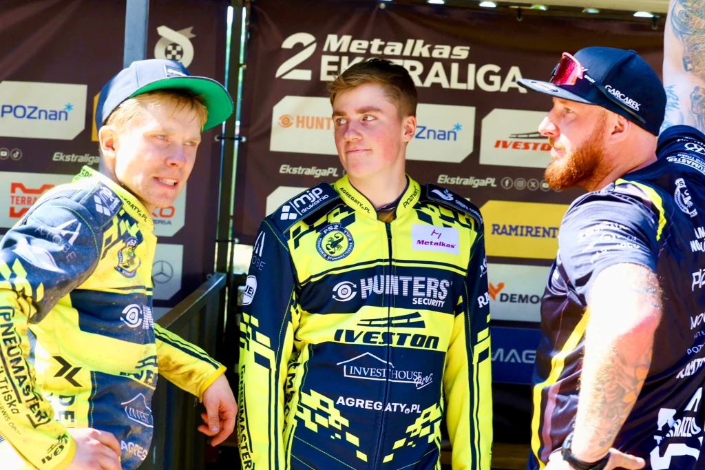 Trzech zawodników drużyny żużlowej w żółto-czarnych kombinezonach rozmawia ze sobą w strefie technicznej z widocznymi logotypami sponsorów na ścianie tła.