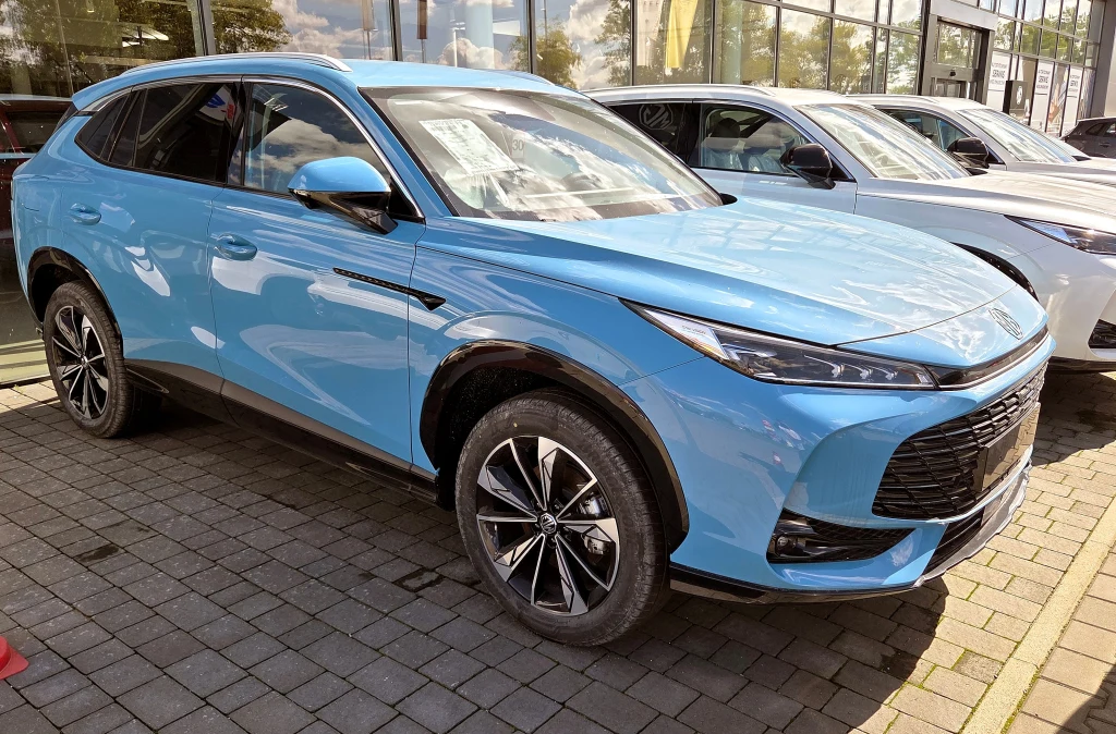 Jasnoniebieski, nowoczesny samochód SUV zaparkowany na chodniku przed salonem samochodowym, lśniąca karoseria, duże felgi, panoramiczne szyby oraz przeszklona przestrzeń wystawowa w tle.