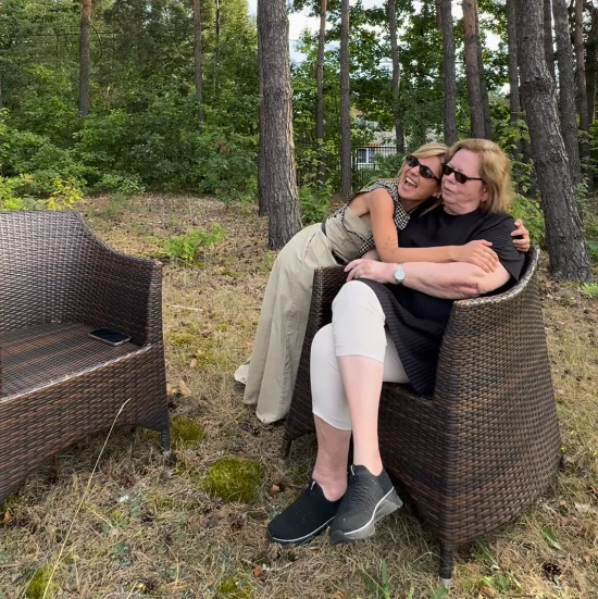 Elżbieta Zapendowska pokazała się na nowym nagraniu z Mimi Wydrzyńską Dwie kobiety siedzą na wiklinowych fotelach w lesie, przytulają się i uśmiechają, otoczone zielenią i drzewami, obok stoi pusty fotel z telefonem na siedzisku.