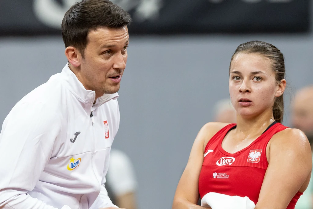 Maja Chwalińska i Dawid Celt Trener w białym dresie rozmawia z zawodniczką w czerwonym stroju sportowym z polskim godłem, która trzyma ręcznik i wygląda na skupioną.
