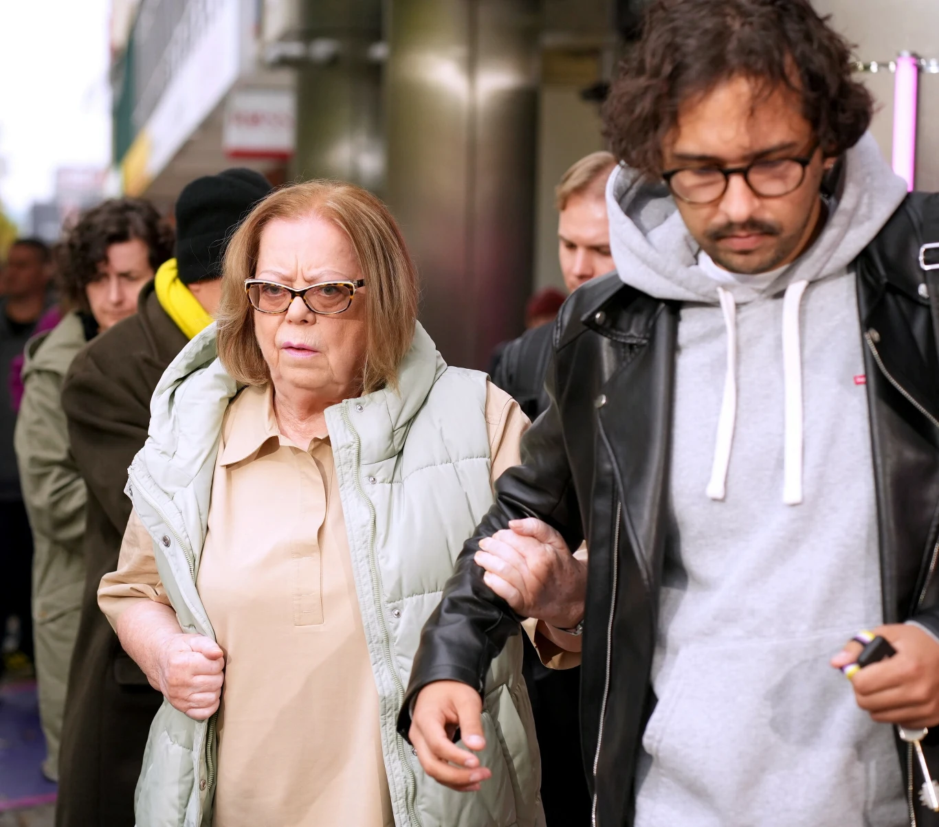 Elżbieta Zapendowska ma poważne problemy ze wzrokiem Starsza kobieta w okularach idzie pod rękę z młodym mężczyzną w skórzanej kurtce i szarej bluzie, w tle widoczne kilka innych osób, scena rozgrywa się na miejskim chodniku.