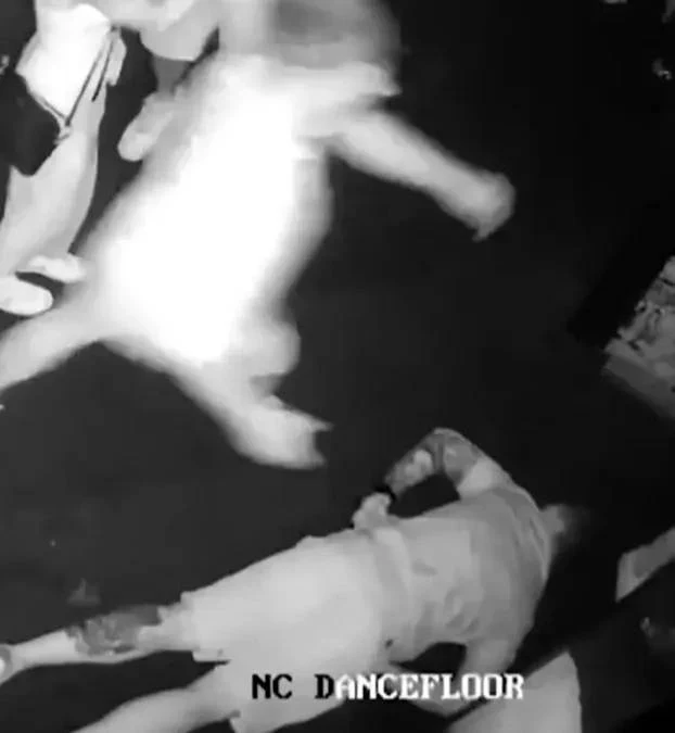 Leżący na podłodze mężczyzna z tatuażami na prawym ramieniu oraz dynamicznie poruszająca się osoba w jasnej sukience, otoczeni przez innych ludzi, scena uchwycona w klubie nocnym na parkiecie.