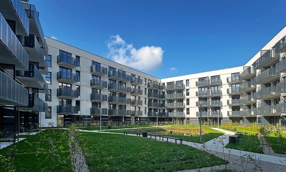 Murapol Portovo Nowoczesny budynek mieszkalny z dużą liczbą balkonów oraz przestronnym zielonym dziedzińcem otoczonym ścieżkami i ławkami, wszystko pod błękitnym niebem z pojedynczą chmurą.